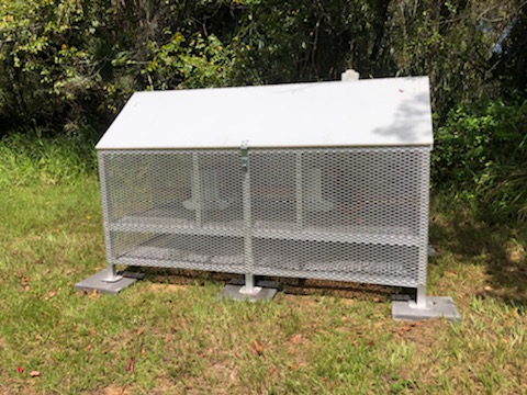 Aluminum framed chicken coop with white roof sitting on pavers in a grassy area near the woods.